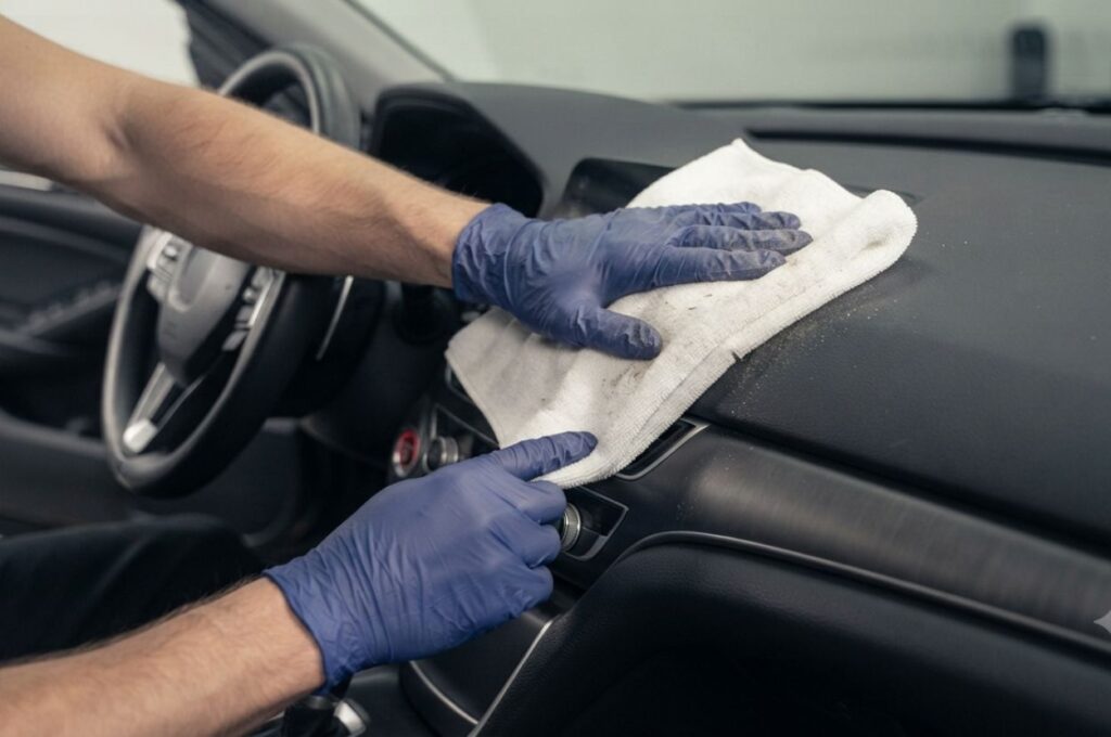 close-up of a detailer wiping a vehicle's dashboard, steering wheel, center console, and door panels with a white microfiber cloth