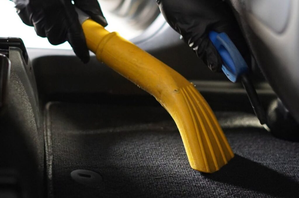 Gloved hands vacuuming a car interior floor mat.